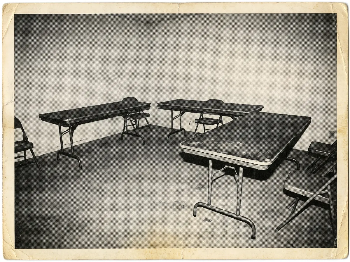 Three folding tables and a few folding chairs in an otherwise empty room, photographed with harsh flash as a vintage press photo