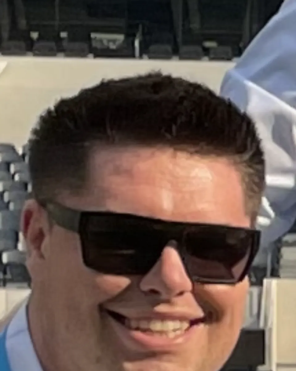 Rob Ercanbrack in sunglasses and a lanyard, smiling in the stands of a professional football stadium, rendered as a vintage newspaper halftone